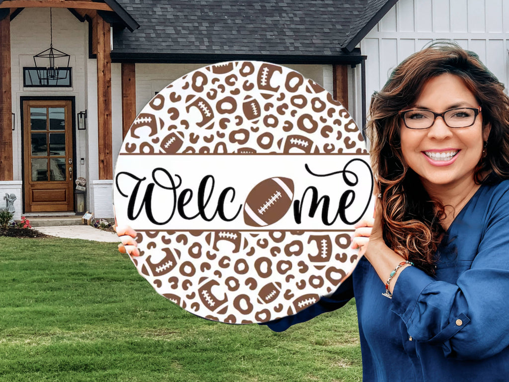 A smiling woman stands in front of a house holding a round sign decorated with footballs and leopard print, featuring the word Welcome with a football replacing the o.