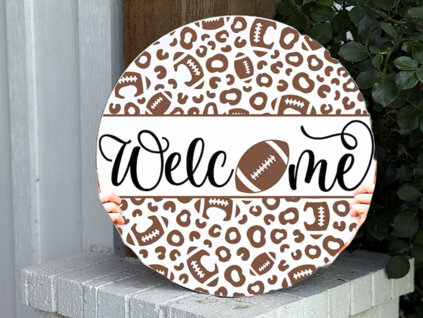 A round sign with a brown and white leopard and football pattern reads “Welcome,” with a football replacing the “o.” Two hands hold the sign, which rests on a white brick ledge next to a green plant.