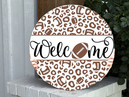 A round sign with a brown and white leopard and football pattern reads “Welcome,” with a football replacing the “o.” Two hands hold the sign, which rests on a white brick ledge next to a green plant.