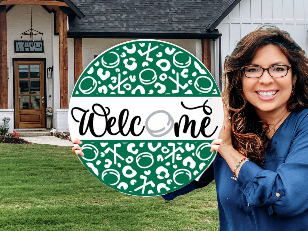 A smiling woman with long brown hair and glasses holds a green and white circular sign that says Welcome in front of a modern house with a wooden entrance and a lawn.