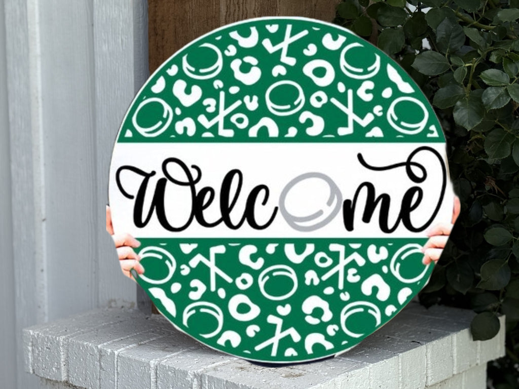 A round green and white welcome sign with abstract patterns is held by two small hands, resting on a white brick surface near a leafy plant and a wooden wall.