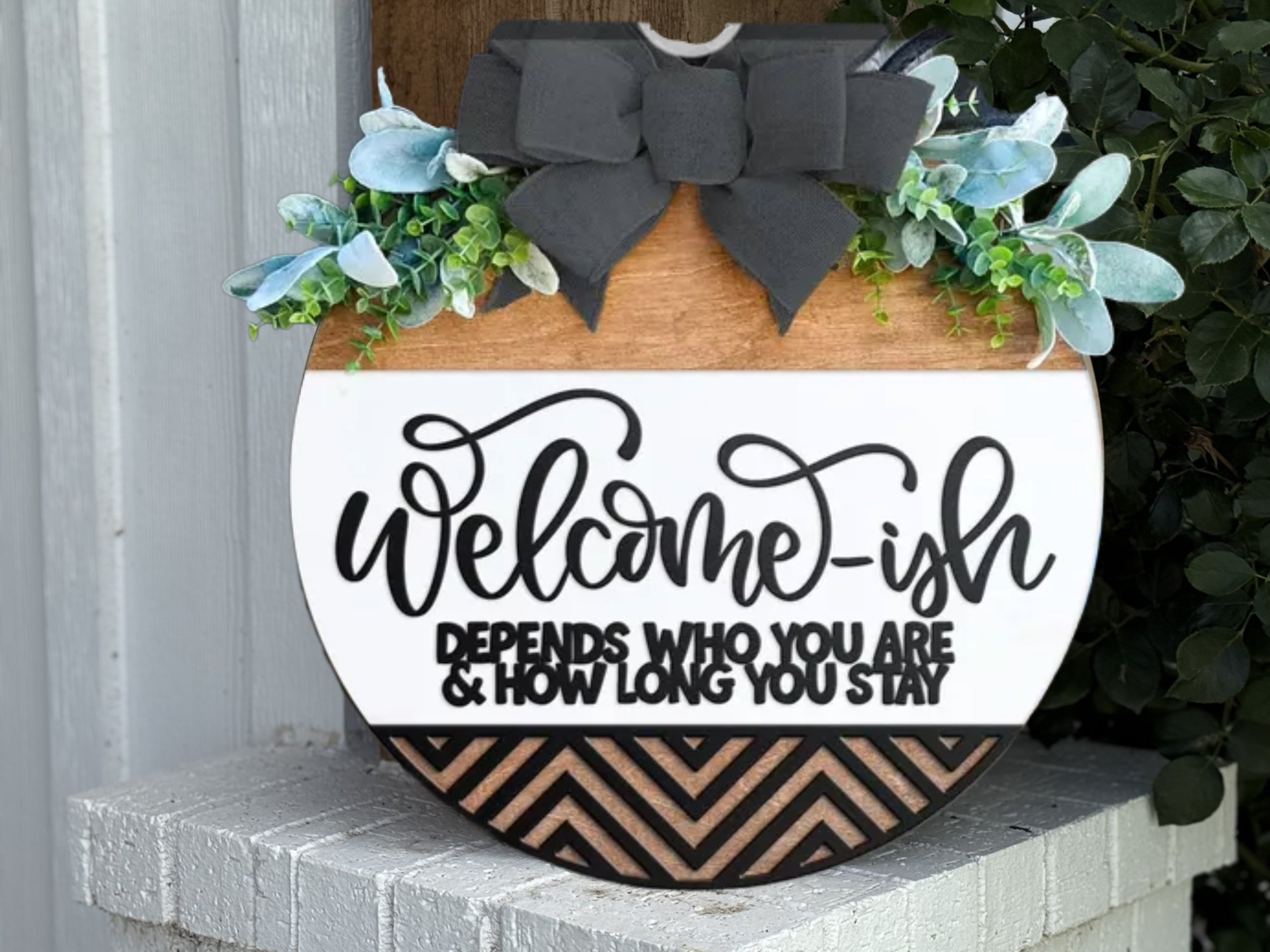 A round wooden sign with greenery and a black bow, reads Welcome-ish, depends who you are & how long you stay, in black text. The bottom features a chevron pattern in black and wood tones.