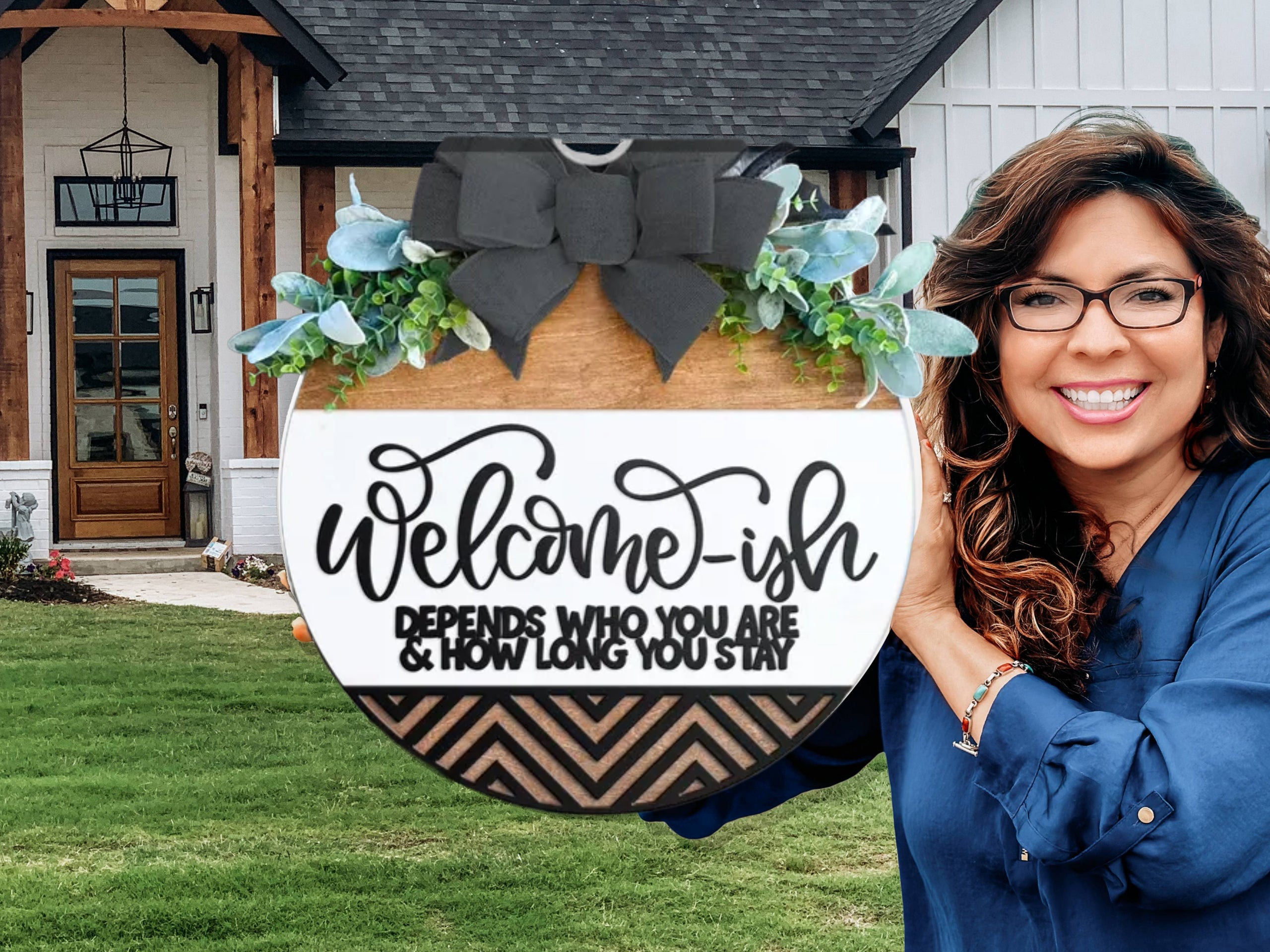 A smiling woman holds a round wooden sign with greenery and a black bow. The sign reads, “Welcome-ish. Depends who you are & how long you stay.” A modern house with a porch is in the background.