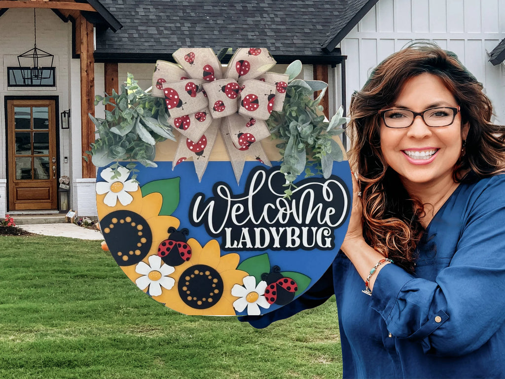 A smiling woman with long, wavy brown hair and glasses holds a colorful “Welcome Ladybug” sign decorated with sunflowers, cartoon ladybugs, green leaves, and a bow with ladybug patterns. She stands outside a white house with wood accents and a green lawn.