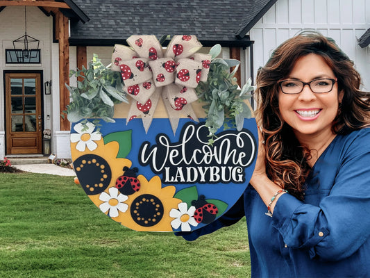 A smiling woman with long, wavy brown hair and glasses holds a colorful “Welcome Ladybug” sign decorated with sunflowers, cartoon ladybugs, green leaves, and a bow with ladybug patterns. She stands outside a white house with wood accents and a green lawn.