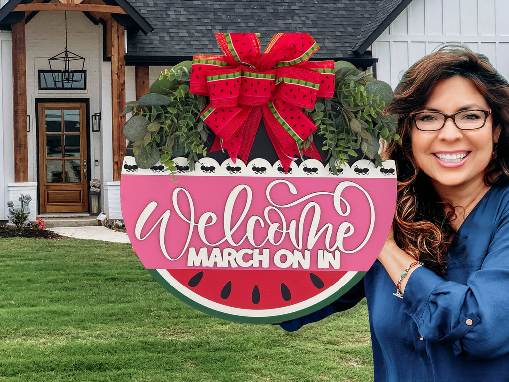 A smiling woman with wavy brown hair and glasses holds a large, round sign shaped like a watermelon slice. The sign is decorated with greenery, a red bow, and reads “Welcome March On In.” She stands in front of a modern house with a green lawn.