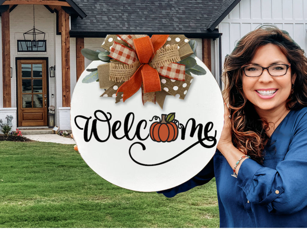 A smiling woman with long, wavy hair and glasses holds a round Welcome sign with a pumpkin design and a decorative bow in front of a modern farmhouse-style home.