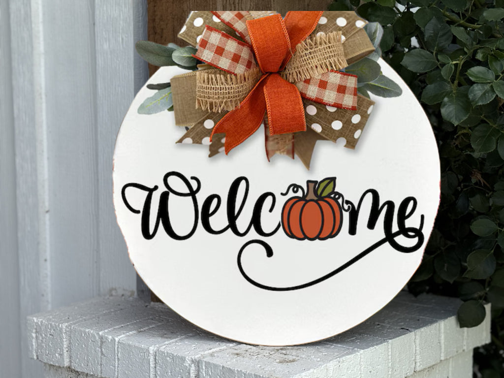 A round white sign with Welcome written in black script, featuring a small pumpkin illustration. The sign is topped with a festive bow made of orange, burlap, gingham, and polka-dot ribbons.