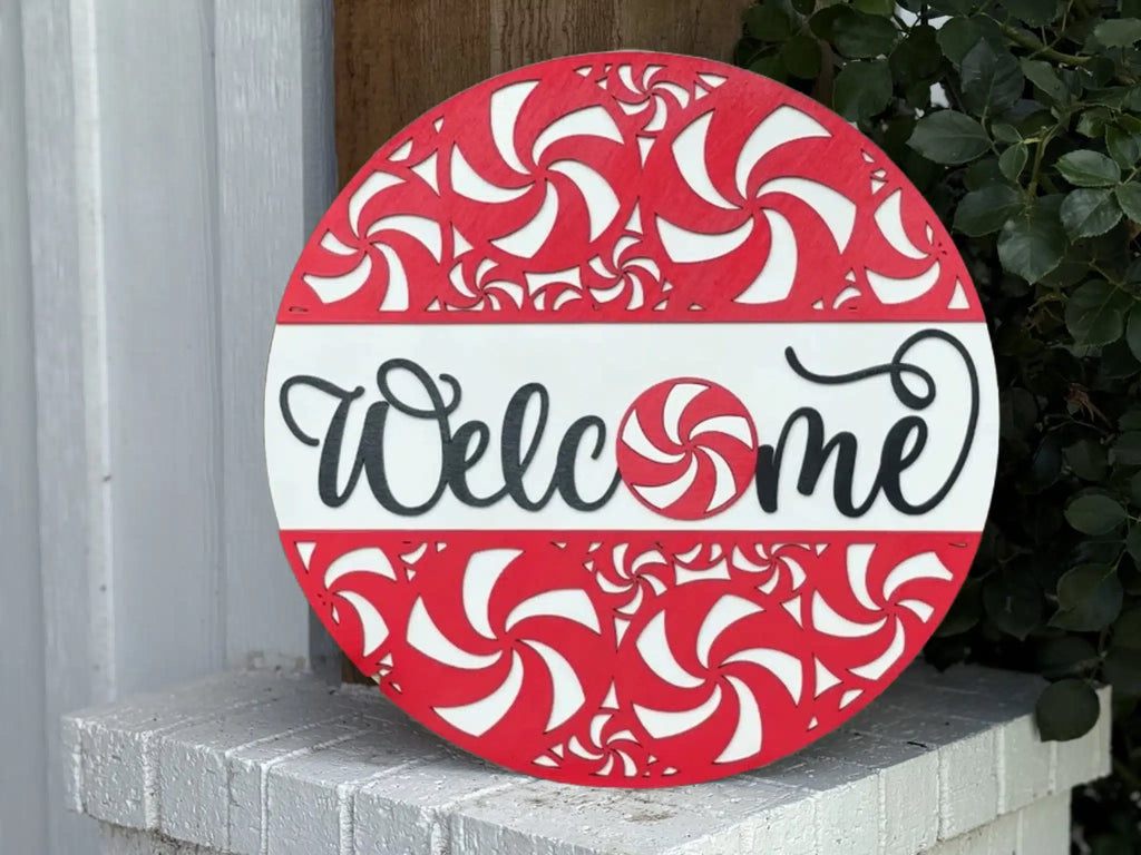 Round sign with red and white peppermint swirl patterns and the word Welcome in black cursive across the center, leaning against a white brick surface next to green leaves.
