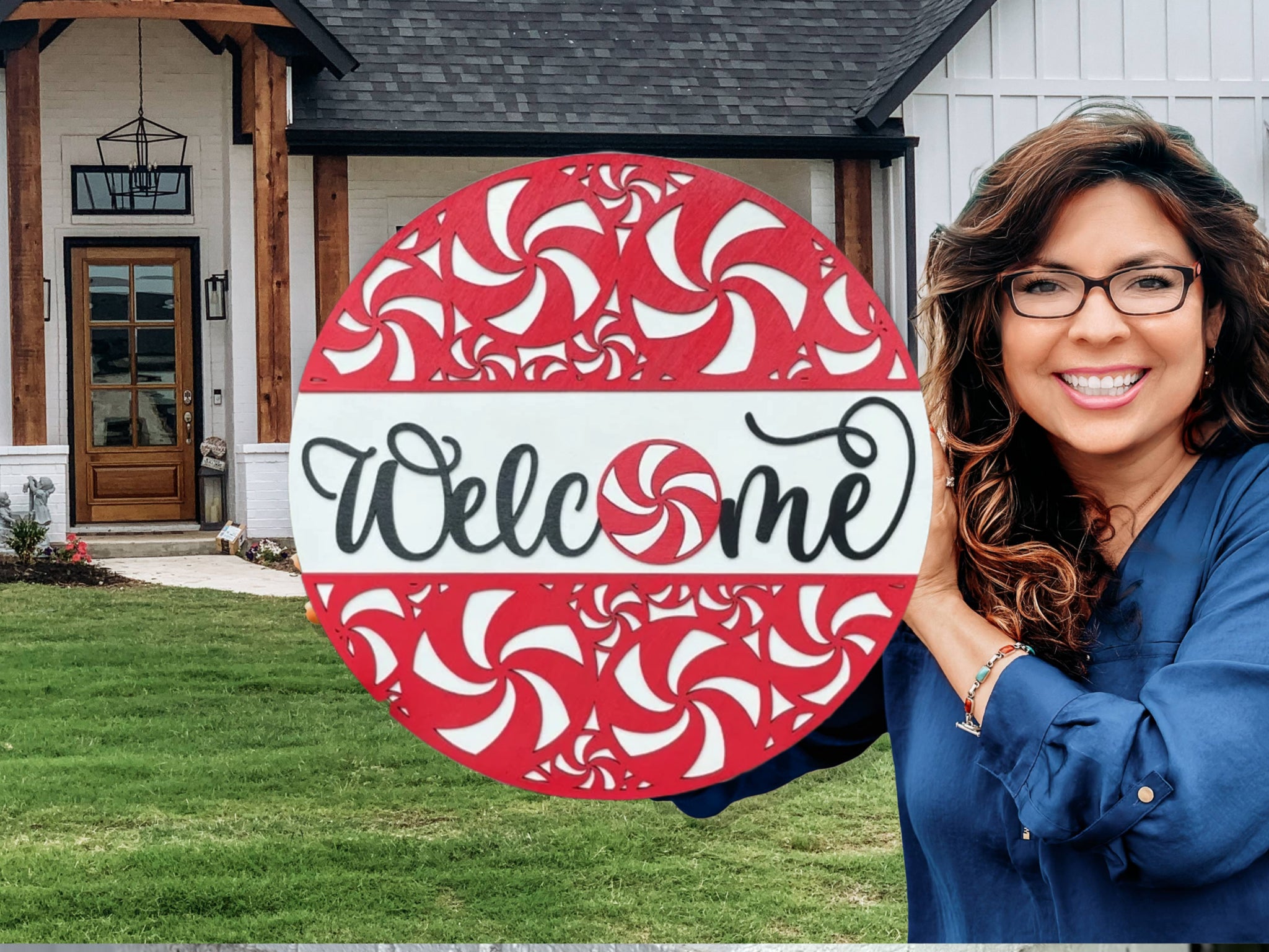 A smiling woman holding a red and white circular Welcome sign with a peppermint pattern, standing in front of a modern house with a lawn and wooden entrance.