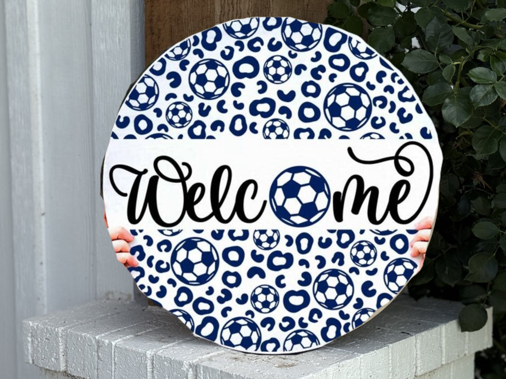 A round sign with blue soccer balls and leopard spots pattern, featuring the word Welcome in cursive, with the o replaced by a soccer ball, held by two hands, placed on a white porch next to green leaves.