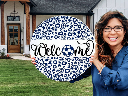 A smiling woman holding a round Welcome sign with blue leopard print and soccer ball designs stands in front of a modern house with a wooden door and green lawn.