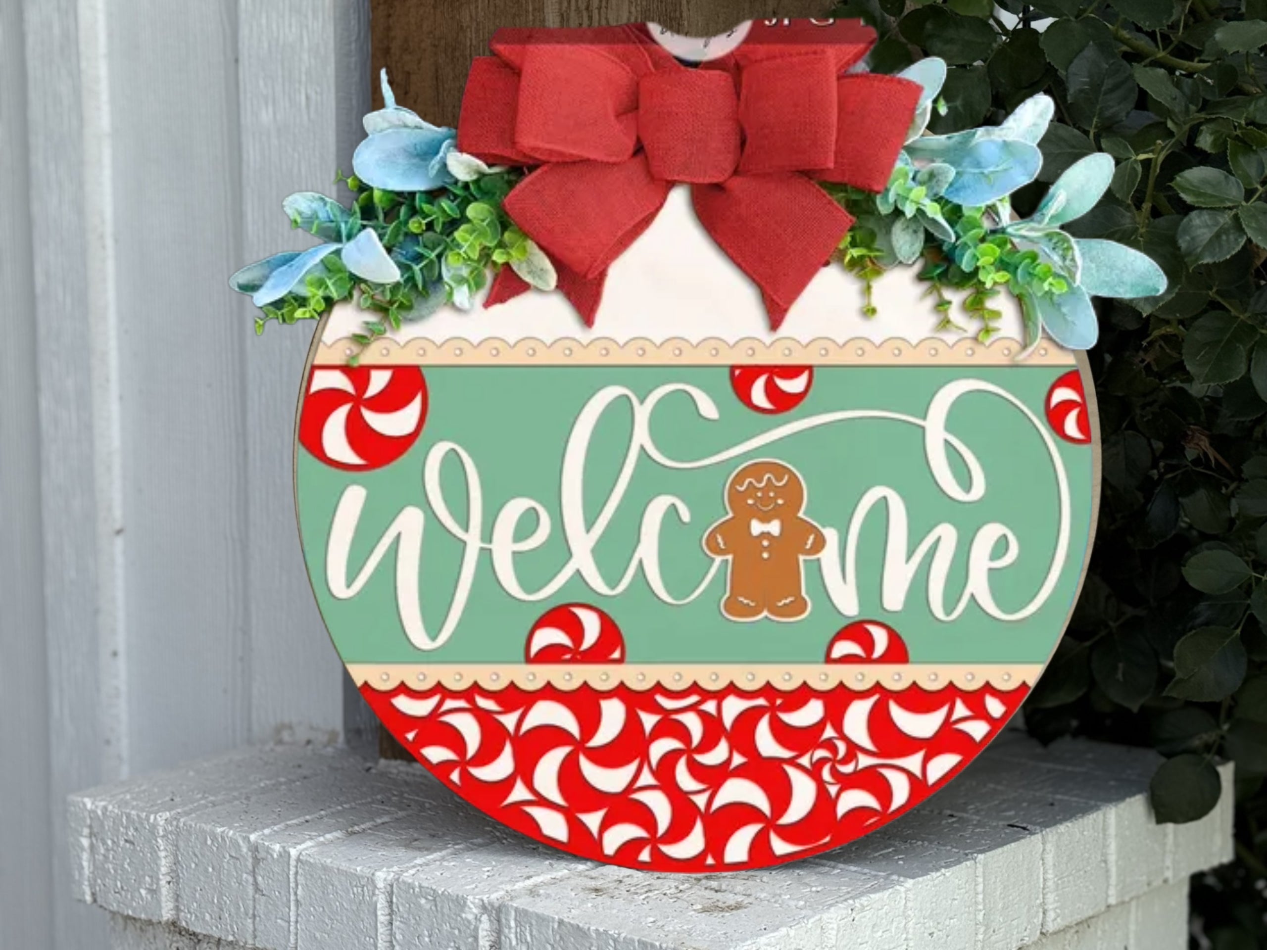 A round holiday welcome sign with a red bow, greenery, and blue flowers on top. The sign reads welcome with a gingerbread figure, and is decorated with red and white peppermint candies.