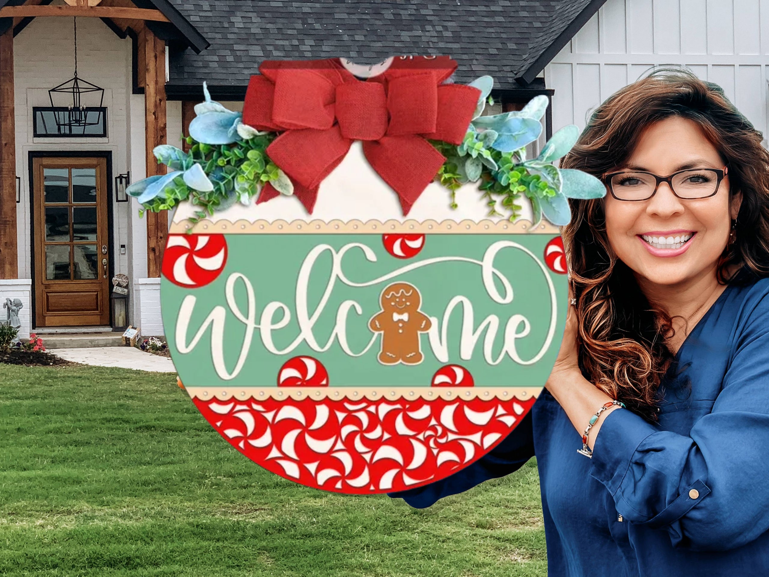 A smiling woman with glasses holds a round, festive Welcome sign decorated with peppermint candies, a gingerbread man, greenery, and a red bow. She stands in front of a modern farmhouse-style home.