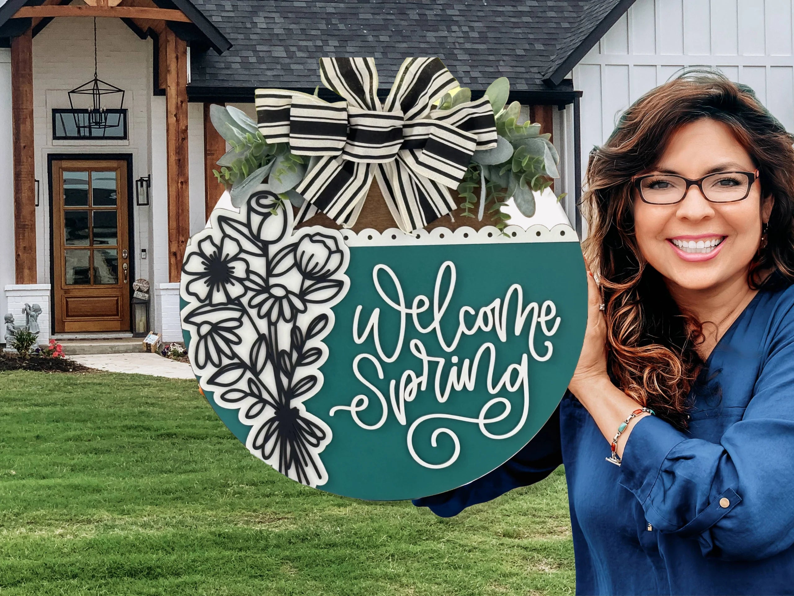 A smiling woman with wavy brown hair and glasses holds a green and teal round sign reading “Welcome Spring” with floral and striped bow decorations, standing in front of a modern farmhouse with a wooden door and manicured lawn.
