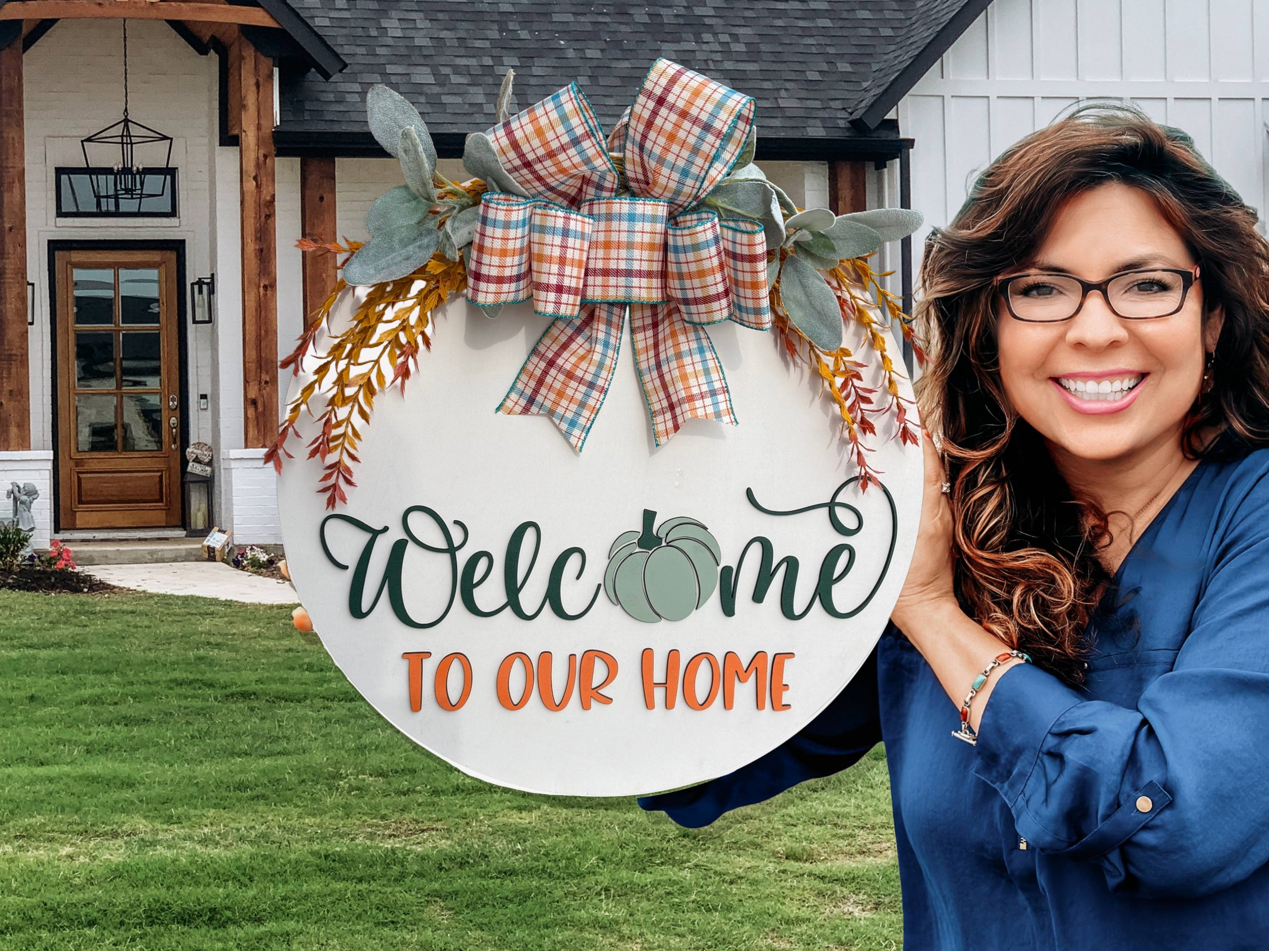 Round white sign with a plaid bow, greenery, and orange sprigs. It reads Welcome to our home, with a green pumpkin in place of the “o” in Welcome. The sign rests on a white brick ledge.