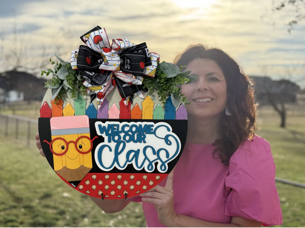 Woman in a pink shirt holds a colorful welcome sign shaped like an apple. The sign is decorated with crayons, an owl, and the words Welcome to Our Class, topped with a bow. She stands outdoors with a cloudy sky and grassy field in the background.