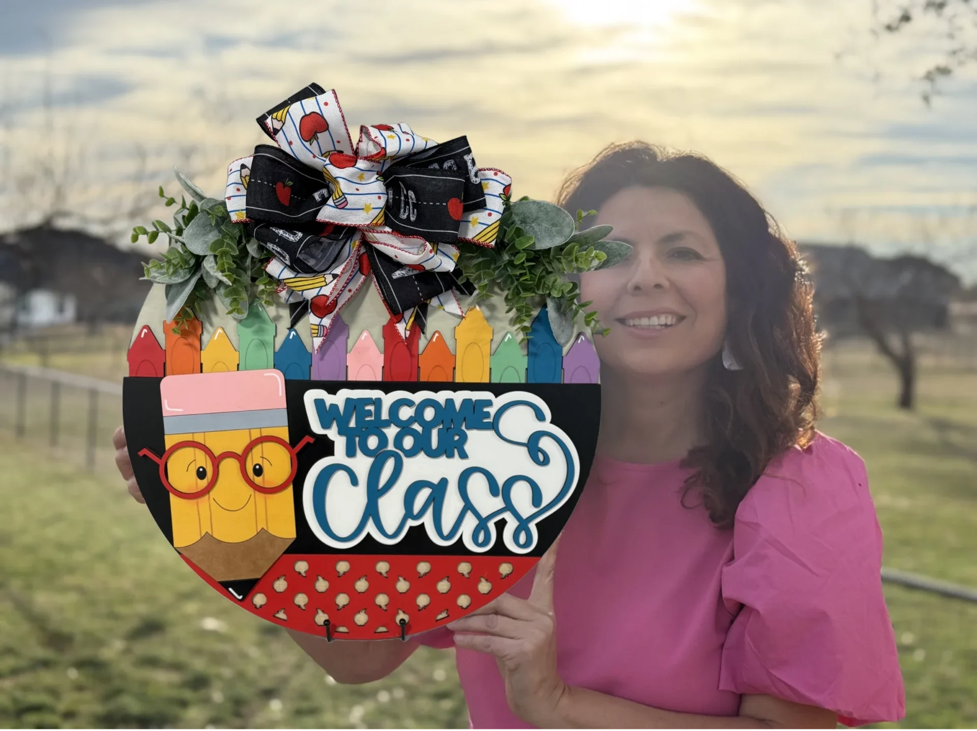 Woman in a pink shirt holds a colorful welcome sign shaped like an apple. The sign is decorated with crayons, an owl, and the words Welcome to Our Class, topped with a bow. She stands outdoors with a cloudy sky and grassy field in the background.
