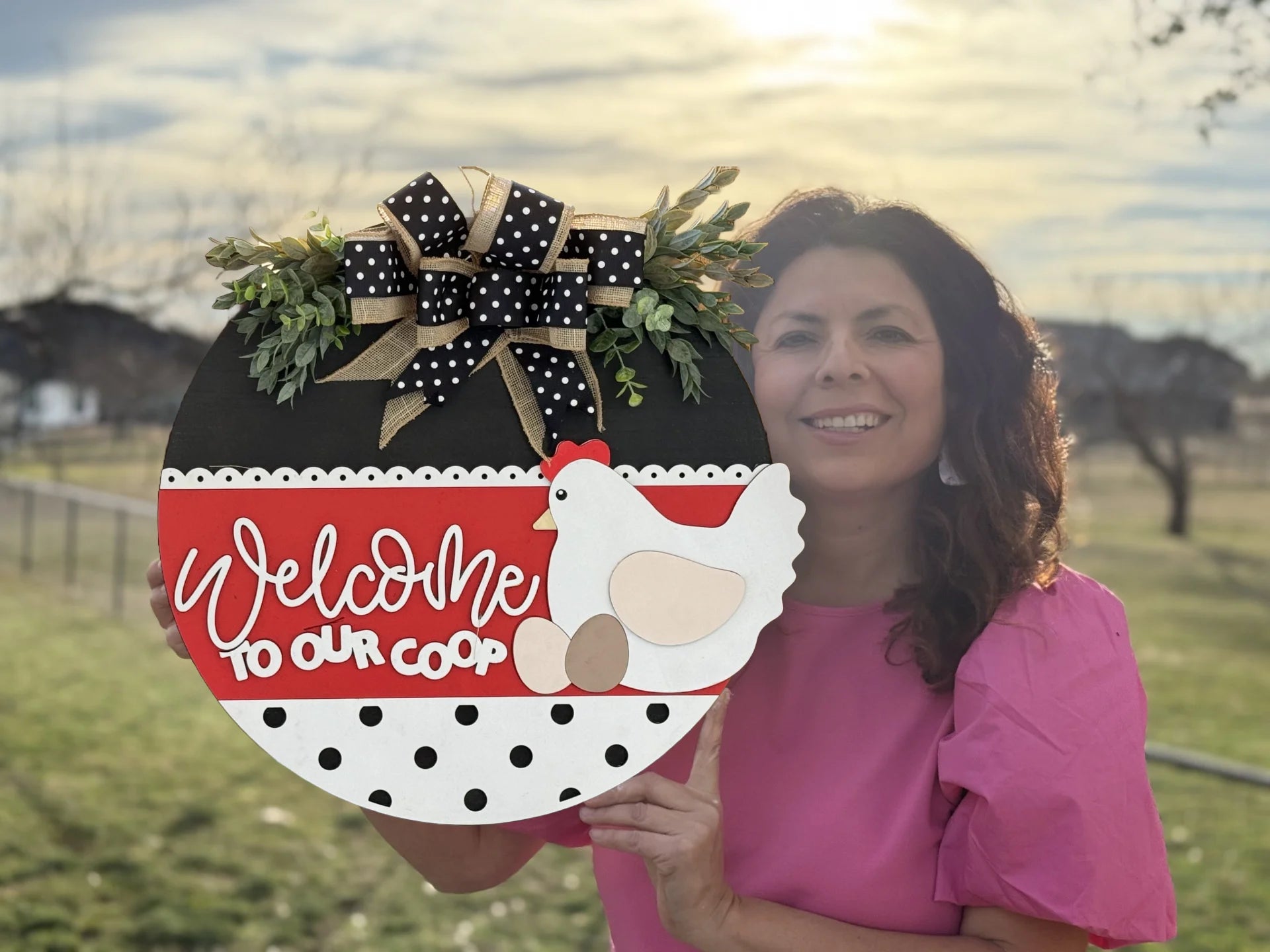 A woman in a pink blouse holds the If Were Not Home Check the Rink decorative door sign featuring a hen, eggs on a polka dot background, and adorned with a black, white, and gold bow with greenery. The scene is set outdoors with grass and trees under a cloudy sky.