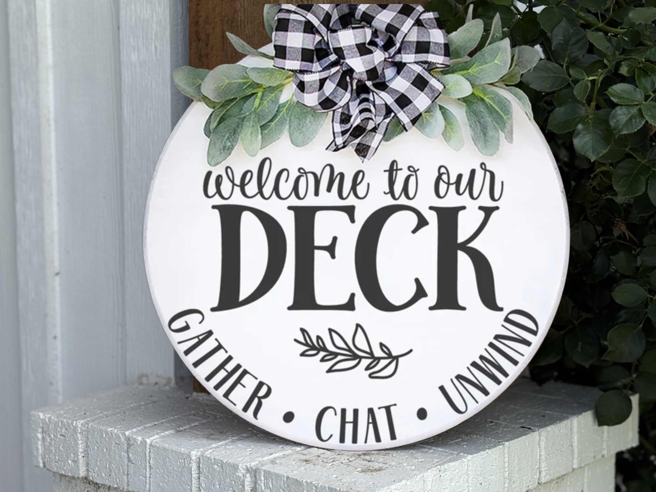 A round white sign with greenery and a black-and-white plaid bow reads “Welcome to our Deck. Gather, Chat, Unwind,” and rests on a white brick ledge near some plants.