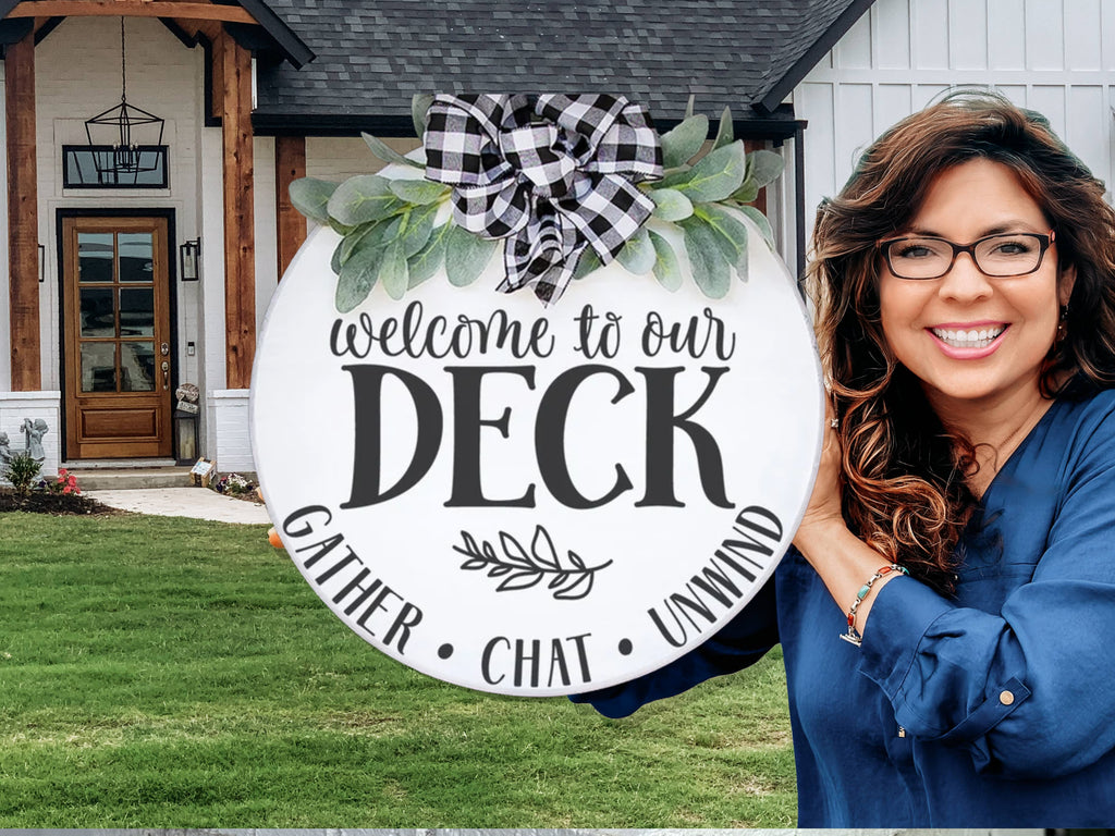 A smiling woman with long brown hair and glasses holds a round sign that reads, Welcome to our deck. Gather. Chat. Unwind. The sign is decorated with greenery and a black-and-white checkered bow. A modern house is in the background.