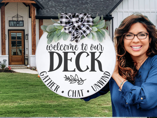 A smiling woman with long brown hair and glasses holds a round sign that reads, Welcome to our deck. Gather. Chat. Unwind. The sign is decorated with greenery and a black-and-white checkered bow. A modern house is in the background.