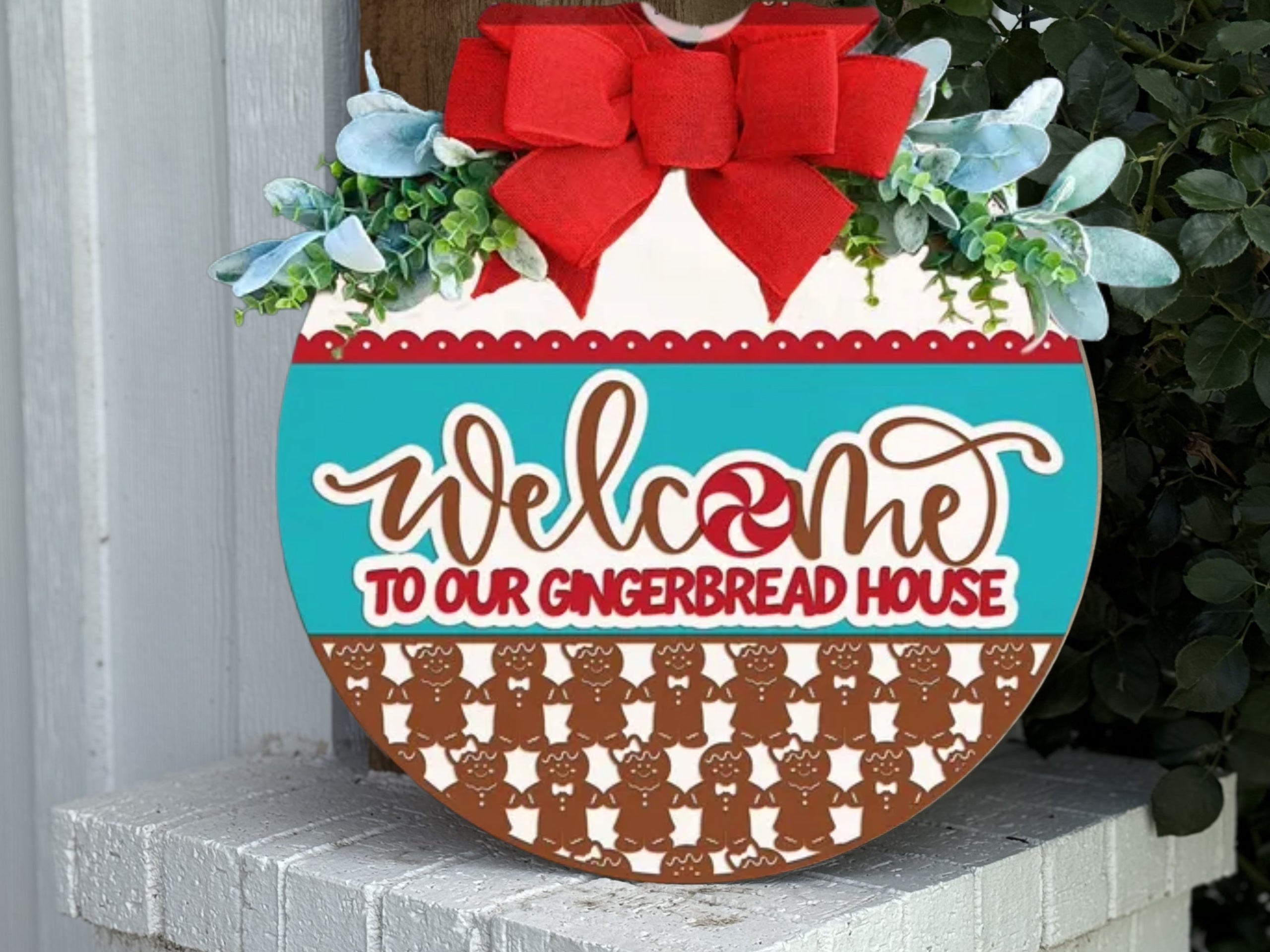 A round sign decorated with greenery and a red bow reads, Welcome to our Gingerbread House in playful lettering, with gingerbread men patterns along the bottom. The sign is displayed on a white brick surface.