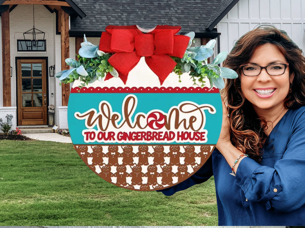 A smiling woman stands outside a house, holding a round sign decorated with a red bow, greenery, and gingerbread men. The sign reads, Welcome to our gingerbread house.