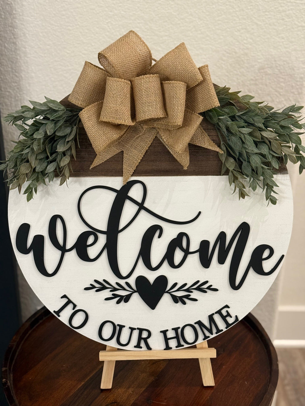 A circular wooden sign with the word Welcome in black cursive, a heart below, and the text To Our Home in block letters. It features a large burlap bow and green foliage on top. The sign rests on a small wooden easel against a white wall.