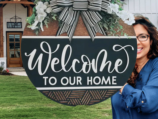 A round wooden sign with white flowers, greenery, and a striped bow reads Welcome to Our Home. A smiling woman in glasses and a blue shirt stands partially visible on the right side. A house and lawn are in the background.