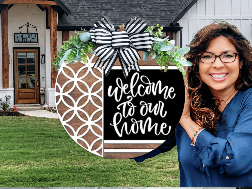 A smiling woman stands outside a house, holding a round decorative sign with a black, white, and wood design that reads “Welcome to our home” and is topped with a striped bow and greenery.