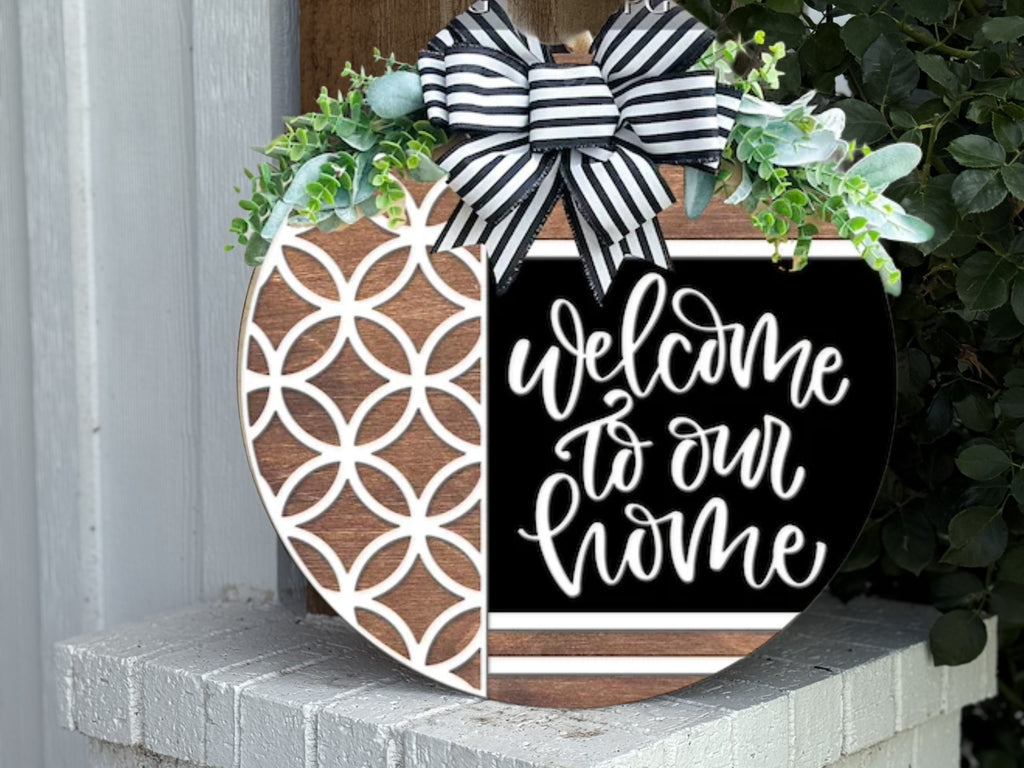 A round wooden sign decorated with greenery and a black-and-white striped bow reads Welcome to our home in white script, with a geometric white pattern on the left side. The sign is displayed outdoors on a white brick ledge.