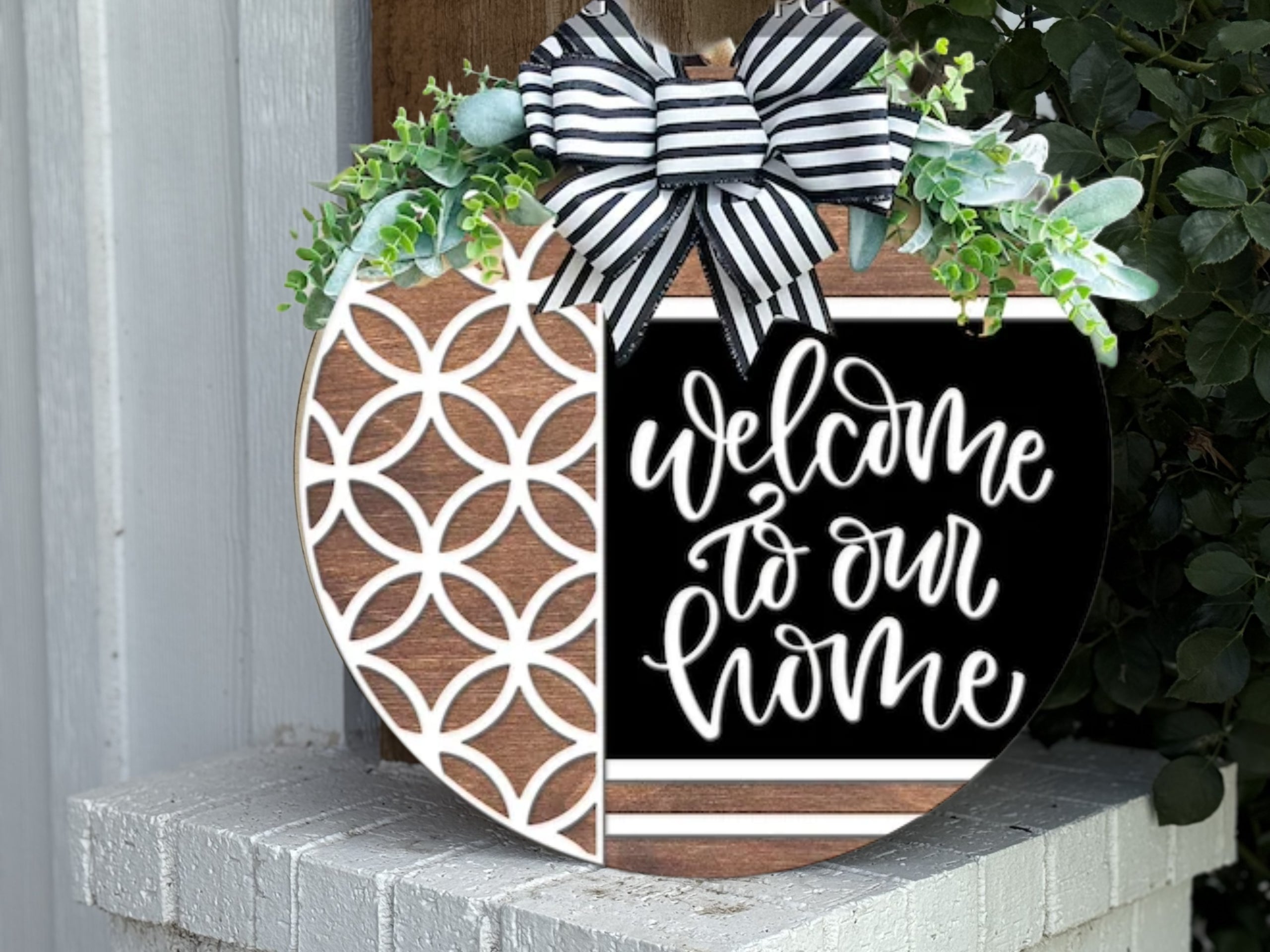 A round wooden sign decorated with greenery and a black-and-white striped bow reads Welcome to our home in white script, with a geometric white pattern on the left side. The sign is displayed outdoors on a white brick ledge.