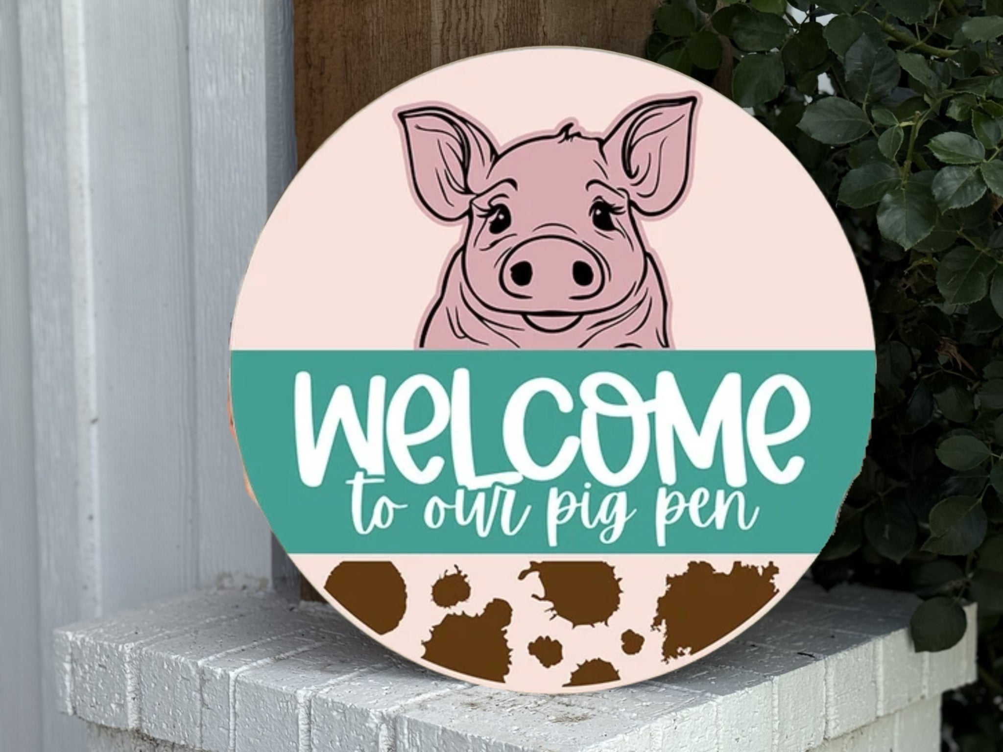 A round sign with a drawing of a pigs face, the text Welcome to our pig pen, and brown pig-shaped paw prints, displayed on a white brick surface near green leaves.