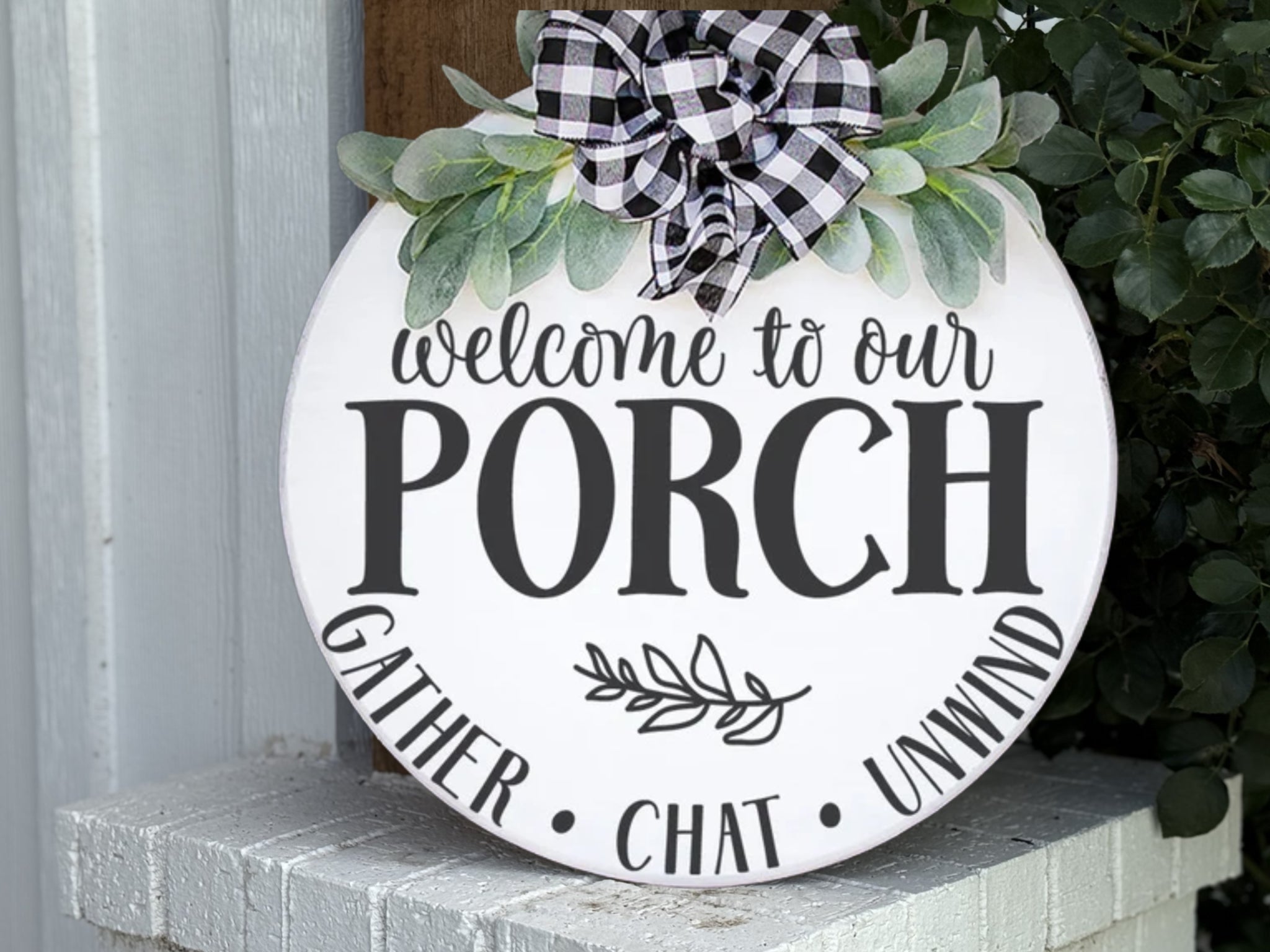 Round white sign with greenery and plaid bow, reads welcome to our PORCH in large letters, and GATHER • CHAT • UNWIND at the bottom; displayed on a white brick surface near leafy plants.