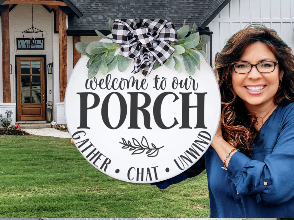 A smiling woman in glasses holds a round sign decorated with greenery and a plaid bow that reads, Welcome to our porch. Gather. Chat. Unwind. She stands outside a modern house with a lawn and porch.