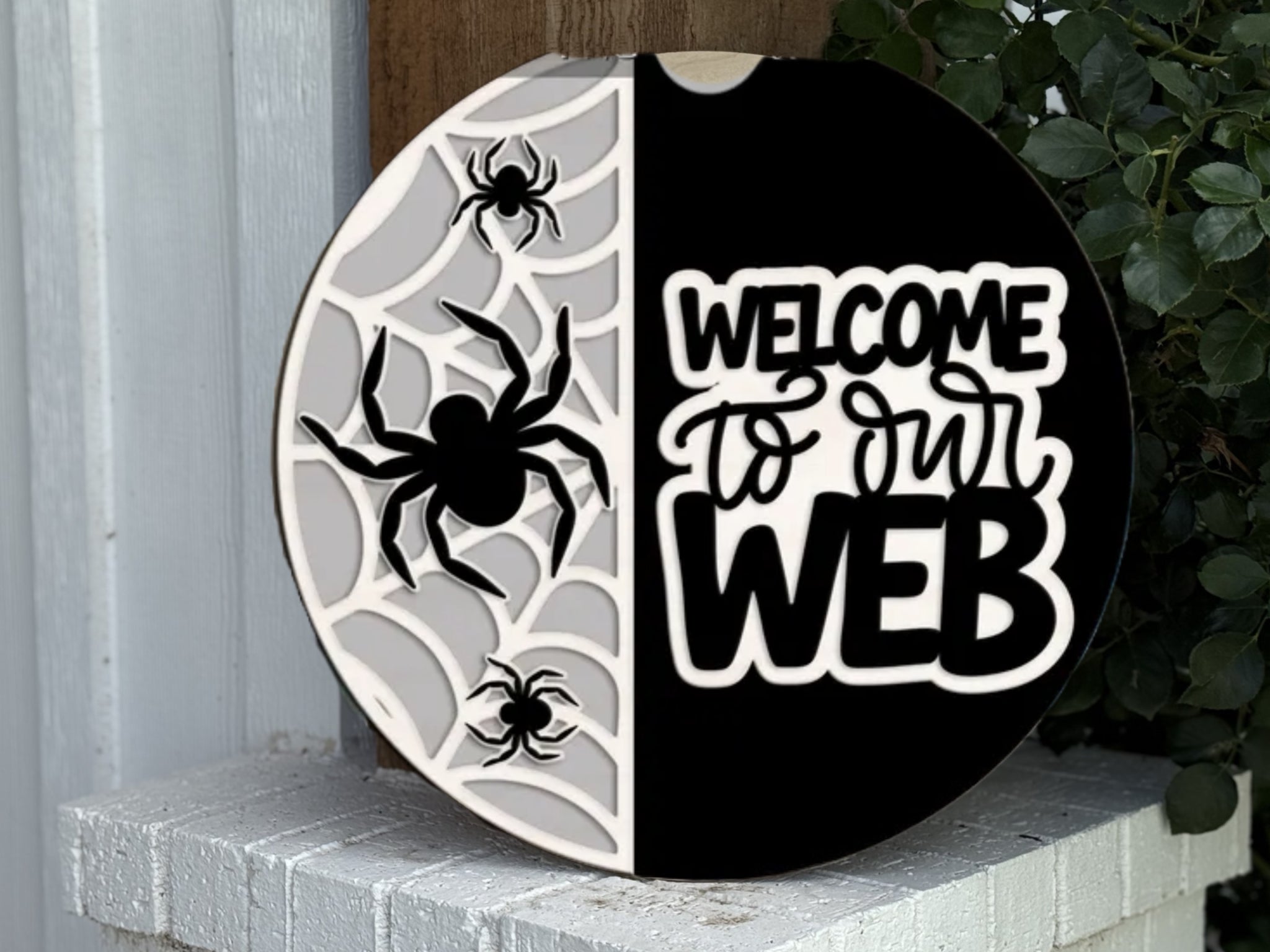 A round sign with black and white sections, featuring three black spiders on a web and the text Welcome to our WEB in bold, playful letters. The sign is displayed on a white brick surface near some green leaves.