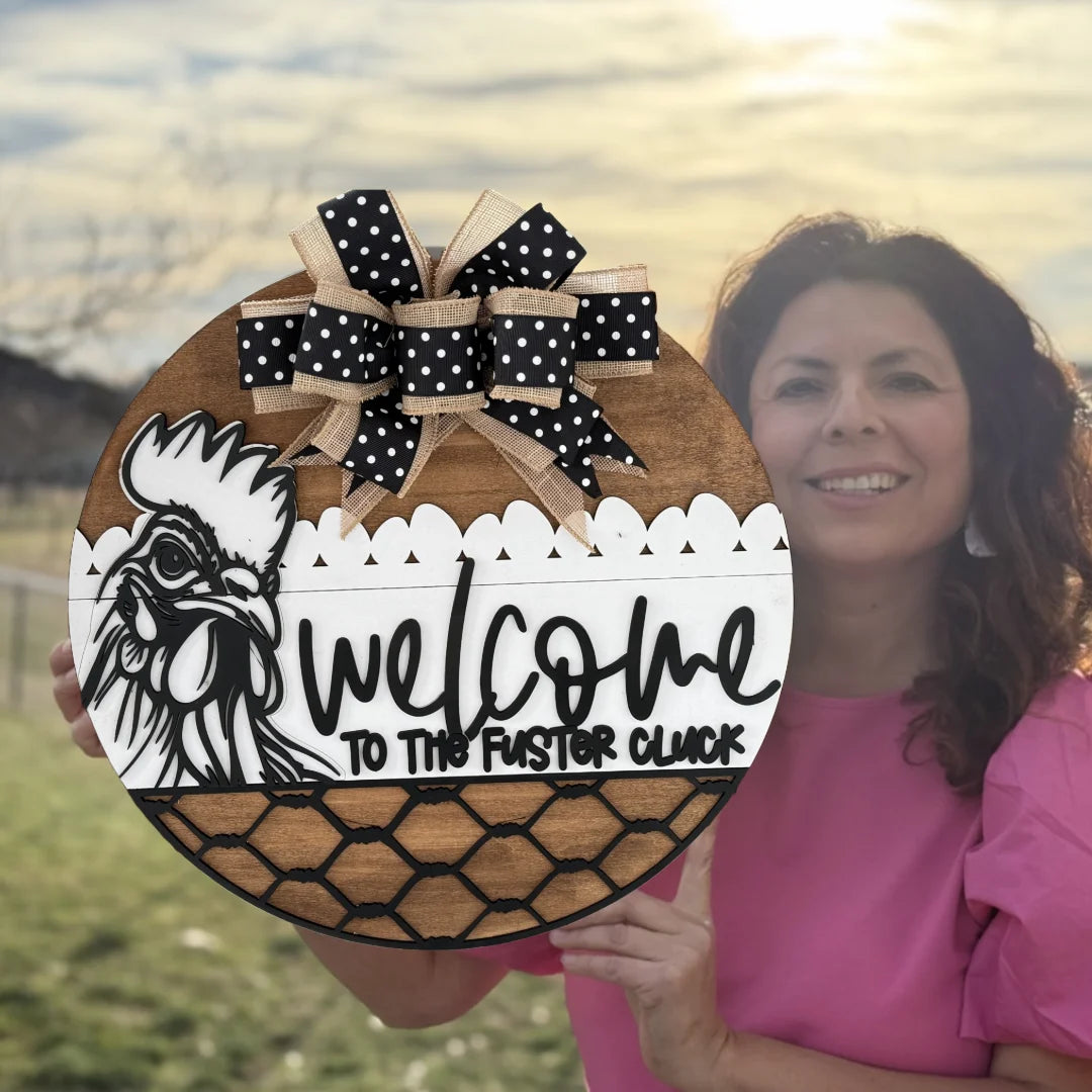 A woman in a pink blouse holds a round wooden sign outdoors. The sign features a rooster illustration, black script text reading Welcome to the Faster Cluck, and a decorative black and beige bow with polka dots. The background shows a blurred grassy field and the sky.
