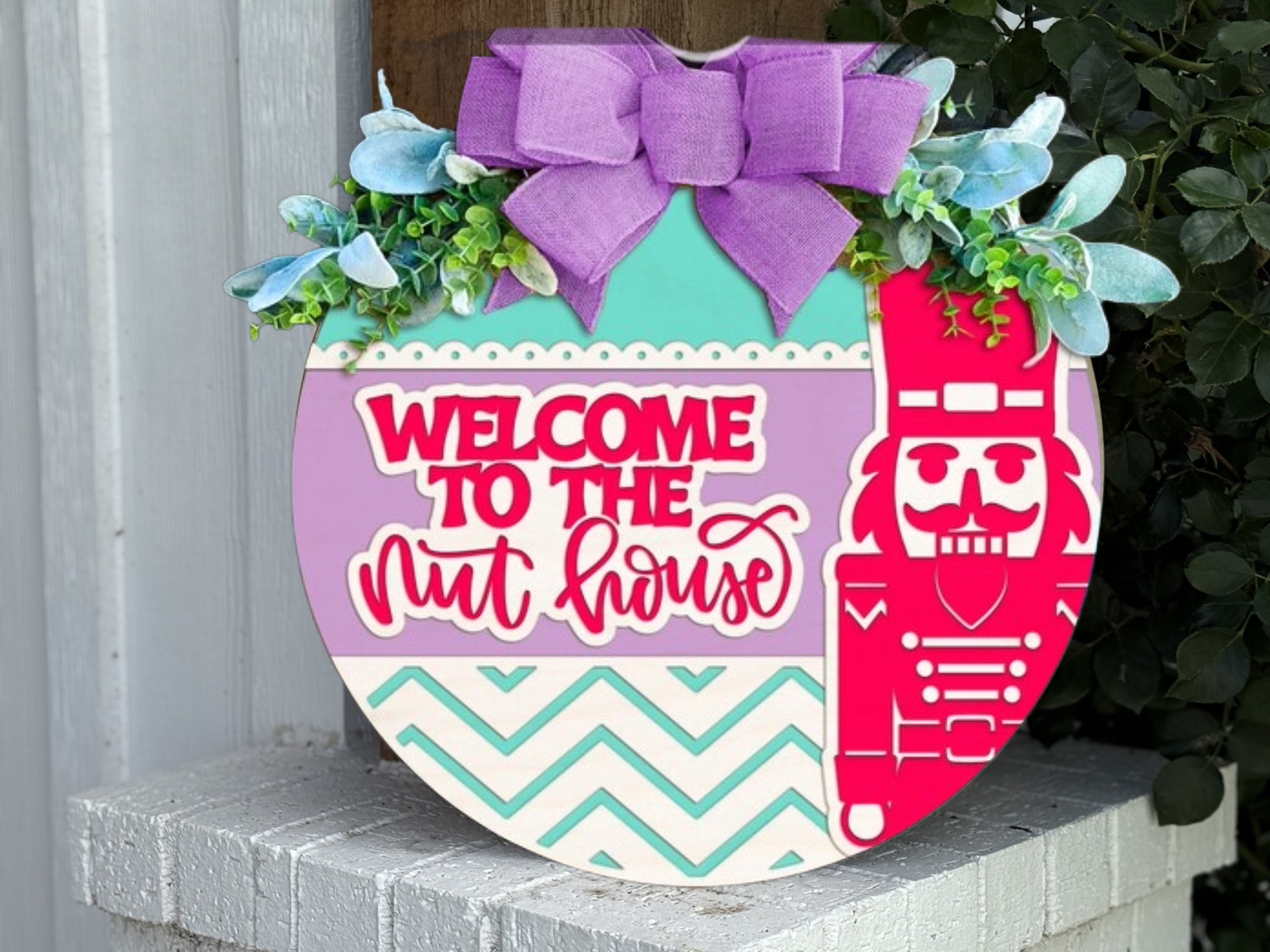 A round decorative sign with a purple bow, greenery, blue flowers, and a design featuring a pink nutcracker. It reads “WELCOME TO THE nut house” in red and white letters on a pastel background.