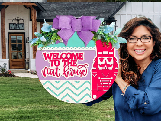 A smiling woman stands outside a house holding a colorful round sign with a nutcracker, a bow, florals, and the text Welcome to the Nut House.