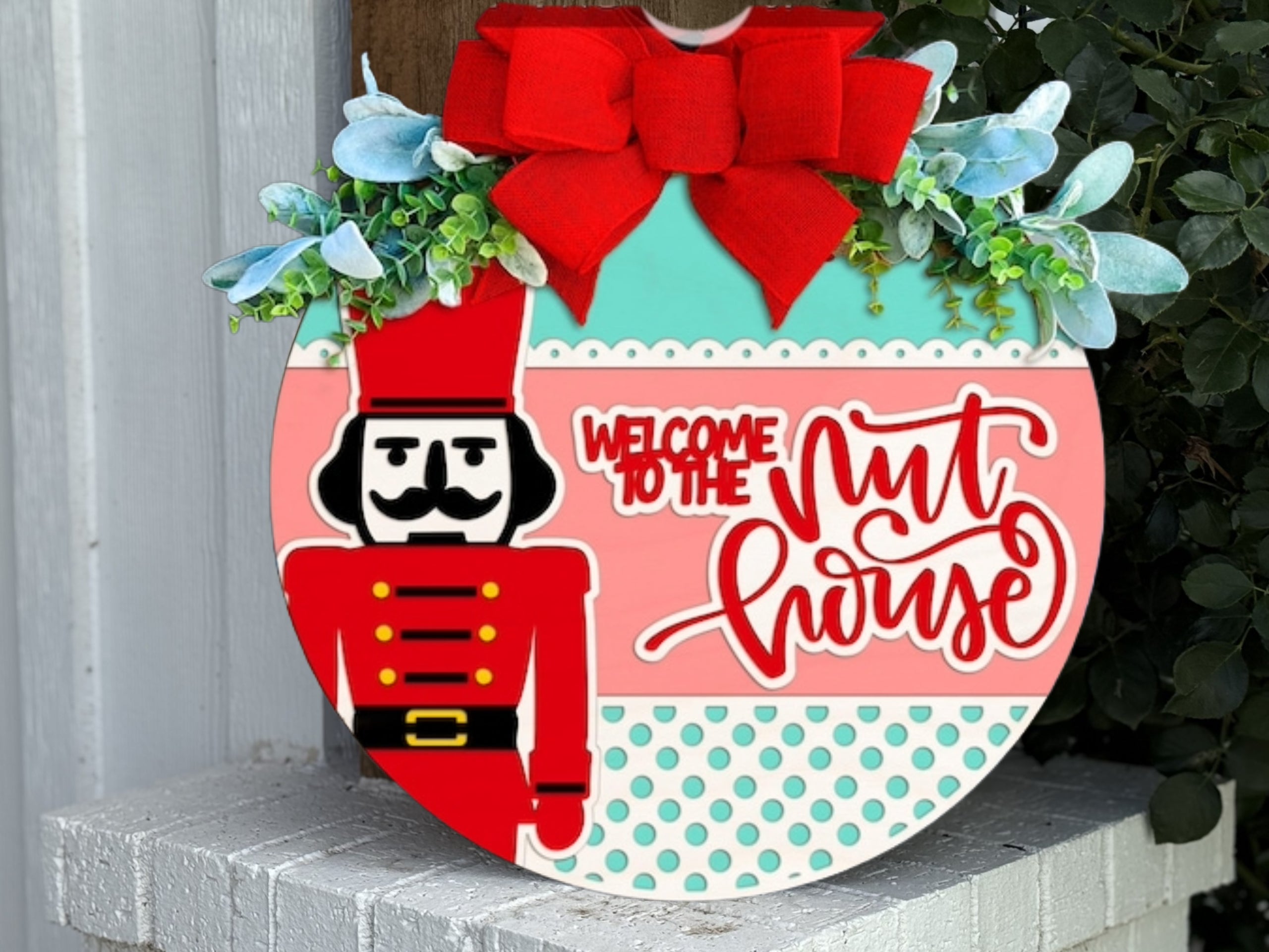 A round door sign decorated with a red bow, greenery, and a nutcracker. The sign features polka dots and stripes and reads, Welcome to the Nut House in playful red and white lettering.