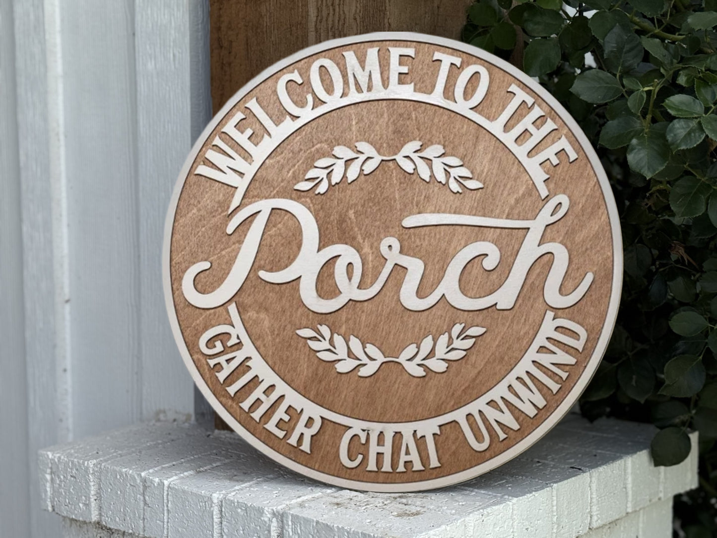 A round wooden sign reads, “Welcome to the Porch. Gather Chat Unwind.” It is leaning against a white brick surface next to green leafy plants.