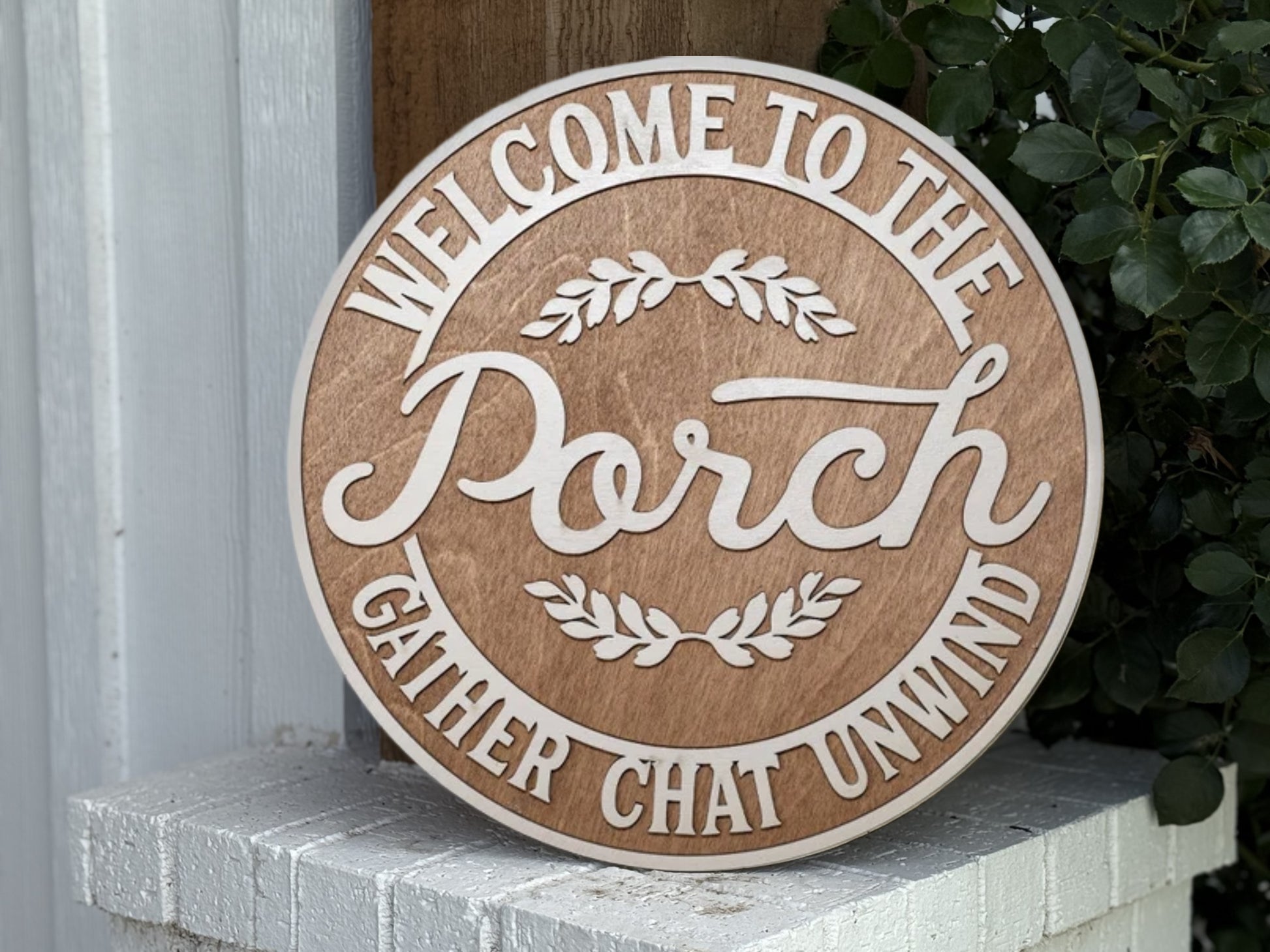 A round wooden sign reads, “Welcome to the Porch. Gather Chat Unwind.” It is leaning against a white brick surface next to green leafy plants.