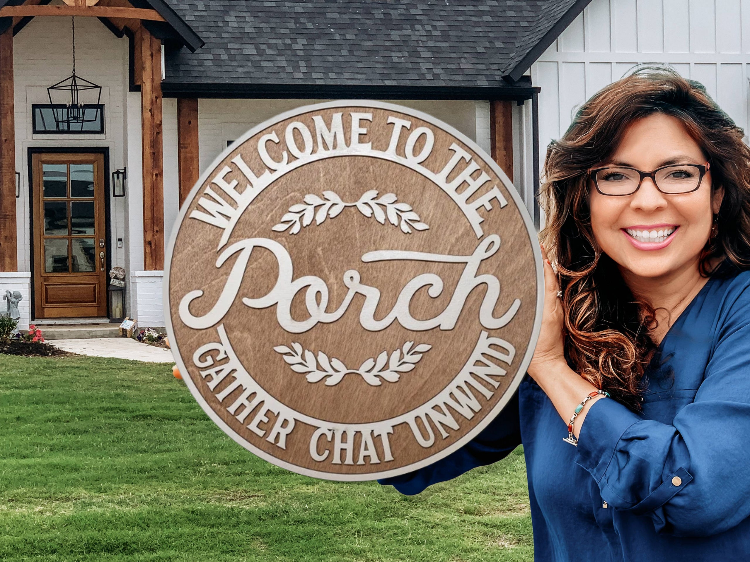 A smiling woman in glasses holds a large round wooden sign that reads, “Welcome to the Porch: Gather, Chat, Unwind,” standing in front of a modern farmhouse-style home with a wooden front door.