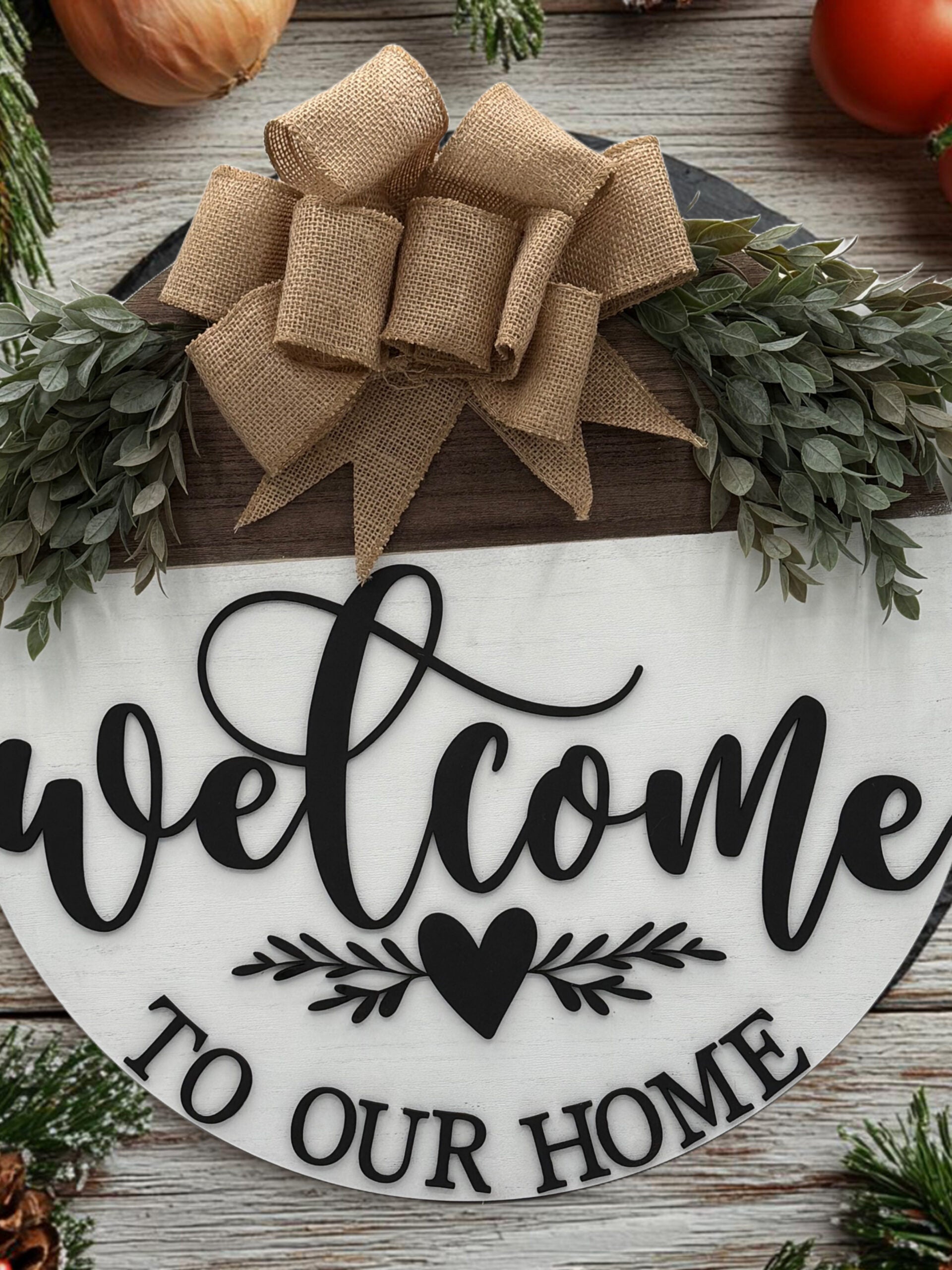 A circular wooden sign with Welcome to Our Home in black script. A heart and floral design are beneath the text. The top is adorned with a burlap bow and green foliage. The background has a rustic wooden texture, with apples and pine branches partially visible around.