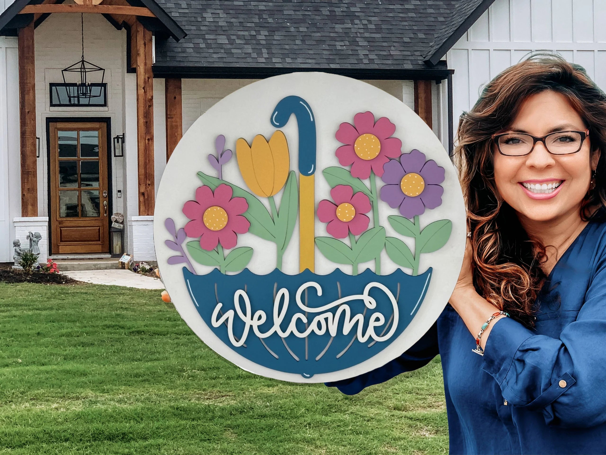 A smiling woman with long brown hair and glasses holds a round sign with colorful flowers in a blue umbrella and the word “welcome.” She stands in front of a modern white house with wood accents, black roof, glass door, and green lawn.