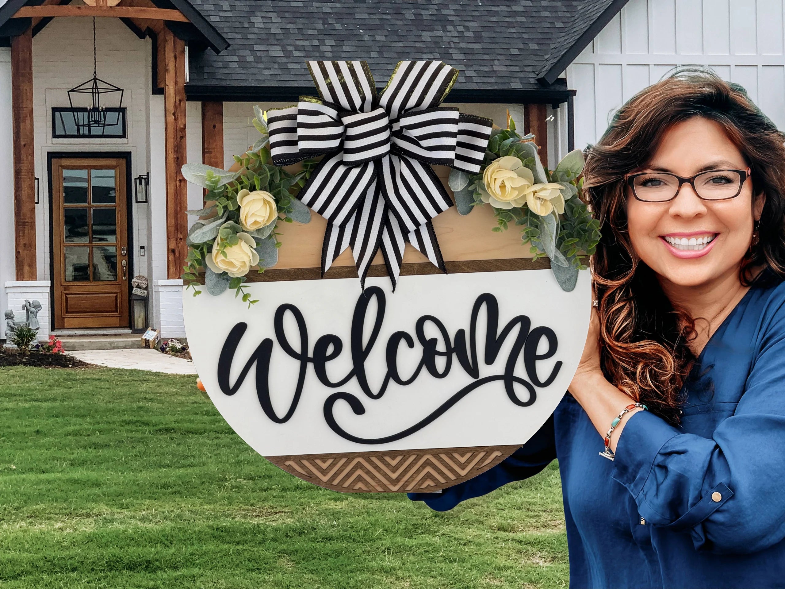 A smiling woman with long brown hair and glasses holds a large round “Welcome” sign decorated with yellow flowers and a black-and-white striped bow. She stands on a green lawn in front of a modern house with wood and white details.