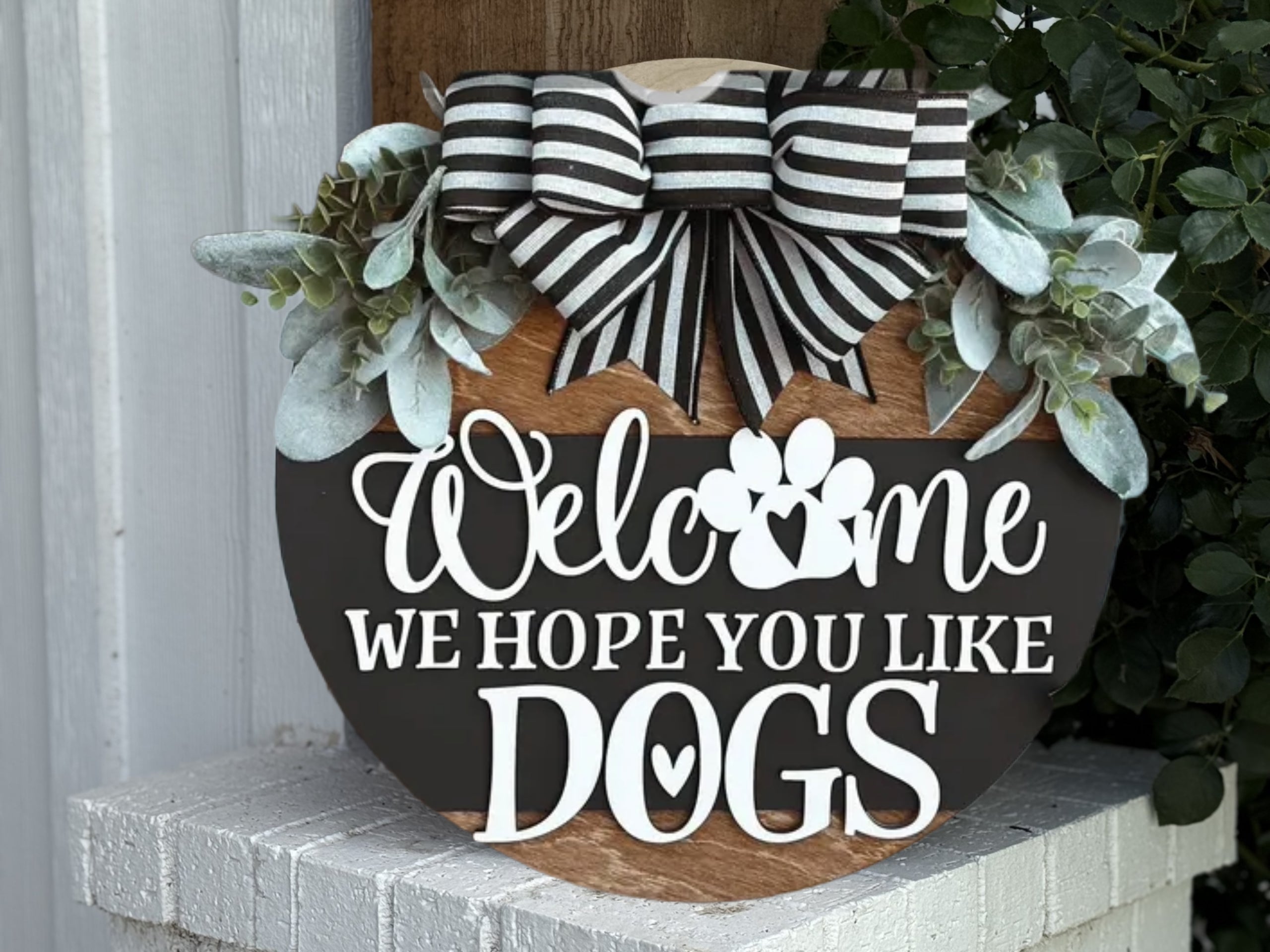 The "Home Is Where the Paws Are" door sign features greenery, a striped bow, and paw print accents. This farmhouse porch decor adds charm to any animal lover's entryway—perfect for dog and cat owners.