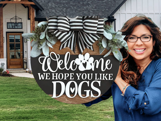 A smiling woman stands outside a house, holding a round sign with a black-and-white striped bow and greenery that reads, “Welcome, we hope you like dogs,” with a paw print replacing the o in Welcome.