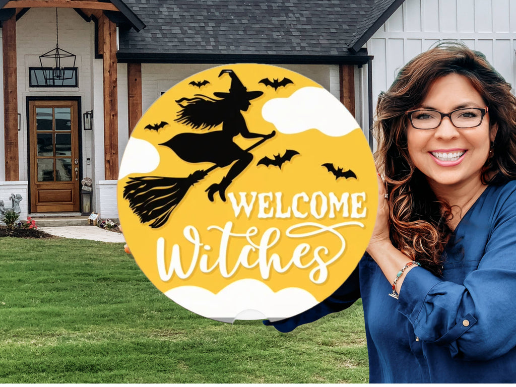 A smiling woman stands outside a house holding a round yellow sign with a silhouette of a witch on a broomstick, bats, clouds, and the words “Welcome Witches” in white script.